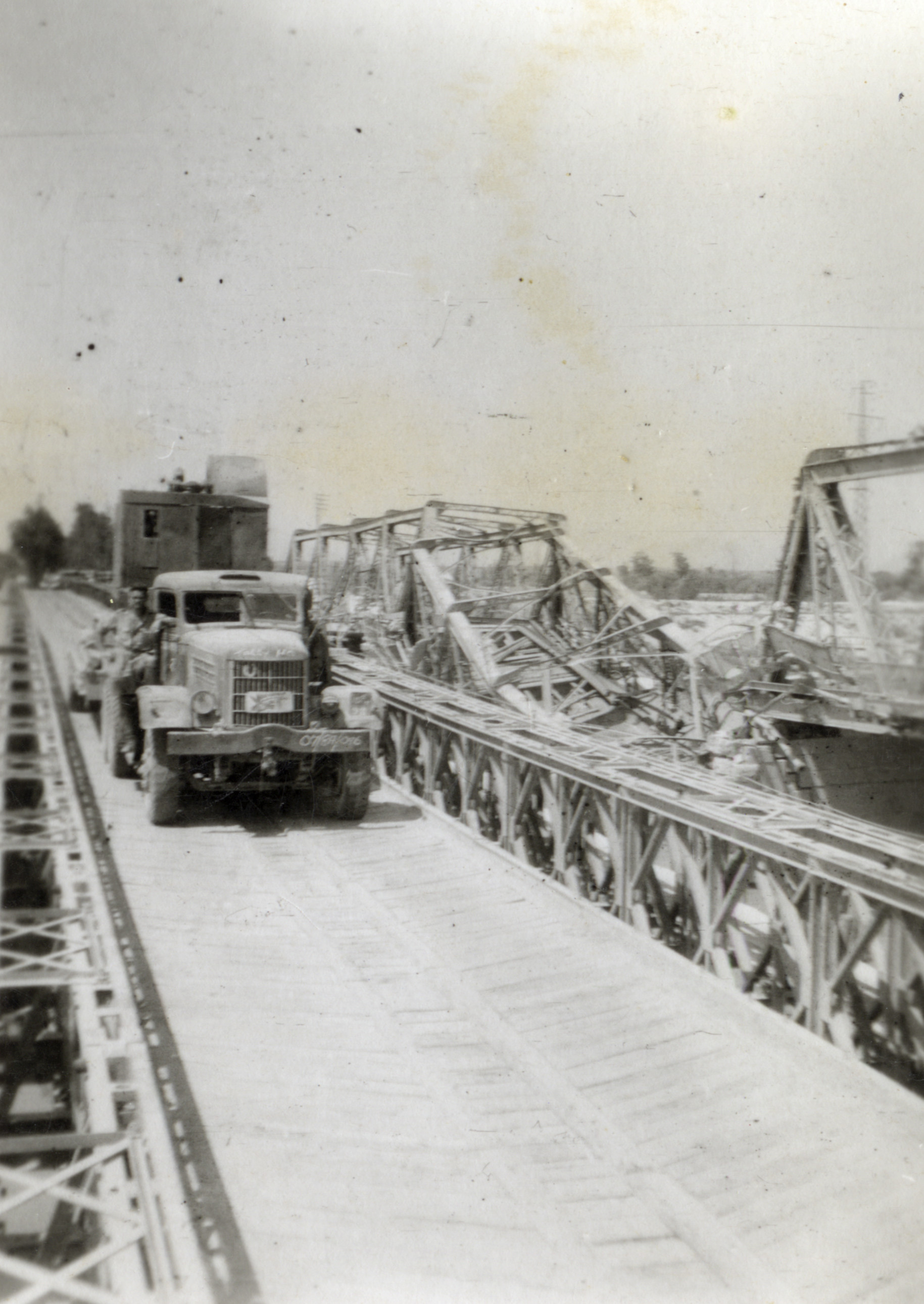 Military Service, World War II; engineers' truck carrying excavator, on Bailey bridge alongside ruined truss bridge.