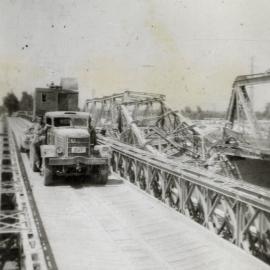 Military Service, World War II; engineers' truck carrying excavator, on Bailey bridge alongside ruined truss bridge.